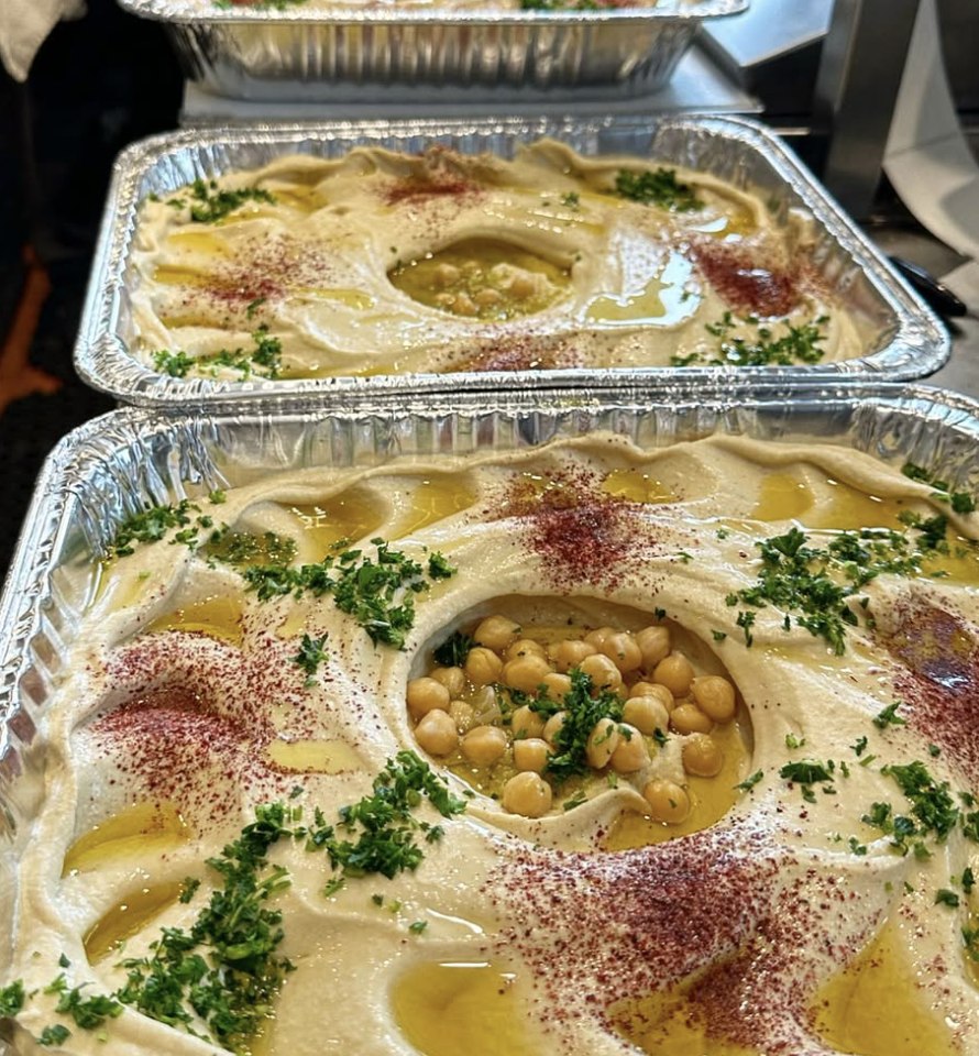A line of full catering trays of hummus, each dressed with olive oil, sumac, parsley and chickpeas